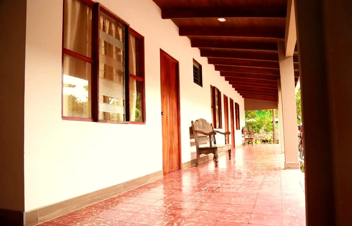 Corredor del hotel con piso de baldosa de barro, paredes blancas, puertas de madera oscura y mecedoras